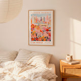 a framed poster of a colorful cityscape with a flower market in the background, hanging on a wall above a bed with striped pillows.