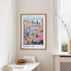 a framed poster of a colorful floral scene hanging on a wall, with a window and a small table with books and a vase in the foreground.