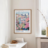 a framed poster of a colorful floral scene hanging on a wall, with a window and a small table with books and a vase in the foreground.