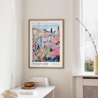 a framed poster of a colorful floral scene hanging on a wall, with a window and a small table with books and a vase in the foreground.