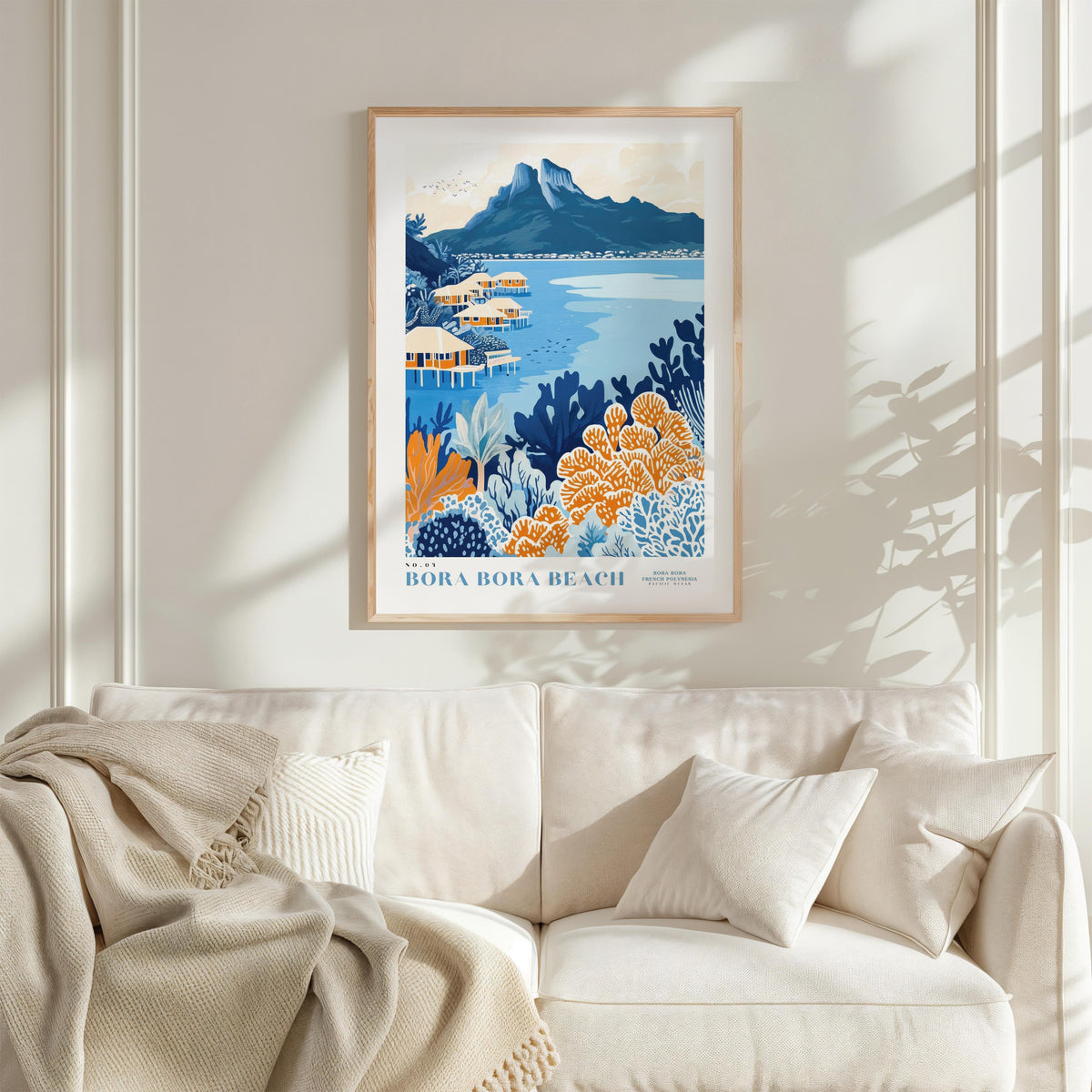 a framed poster of a tropical beach scene with mountains in the background, hanging on a wall above a white couch with a blanket draped over it.