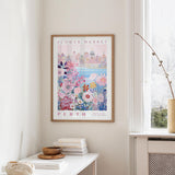 a framed poster of a cityscape with a floral design, hanging on a wall in a room with a window and a small table with books and a bowl on it.