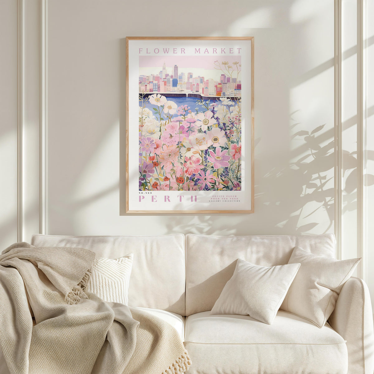 a framed poster of a flower market in Perth, with a cityscape in the background. The poster is hanging on a white wall above a white couch, which has a blanket draped over it.