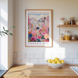 a kitchen scene with a framed poster of a colorful floral cityscape hanging on the wall above a wooden countertop. On the countertop, there is a bowl of lemons and a plant.