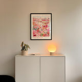 a white cabinet with a potted plant and a lamp on top, set against a white wall. Above the cabinet, there is a framed poster of a flower market.