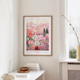 a framed poster of a pink and white floral scene hanging on a wall, with a white table and a vase of flowers nearby.