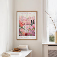 a framed poster of a pink and white floral scene hanging on a wall, with a white table and a vase of flowers nearby.