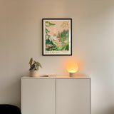 a white cabinet with a plant and a lamp on top, and a framed poster of a tropical landscape hanging on the wall above it.