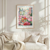 a framed poster of a flower market, with a cityscape in the background. The poster is hanging on a wall above a white couch, which has a blanket draped over it.