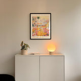 a white cabinet with a potted plant and a lamp on top, set against a white wall. Above the cabinet, there is a framed poster with a colorful, abstract design.