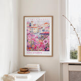 a framed poster of a colorful floral scene hanging on a wall, with a white table and a vase of flowers nearby.