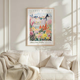 a framed poster of a cityscape with a flower market in the background, hanging on a wall above a white couch with a blanket draped over it.