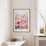 a framed poster of a colorful floral scene hanging on a wall, with a window and a small table with books and a vase in the foreground.