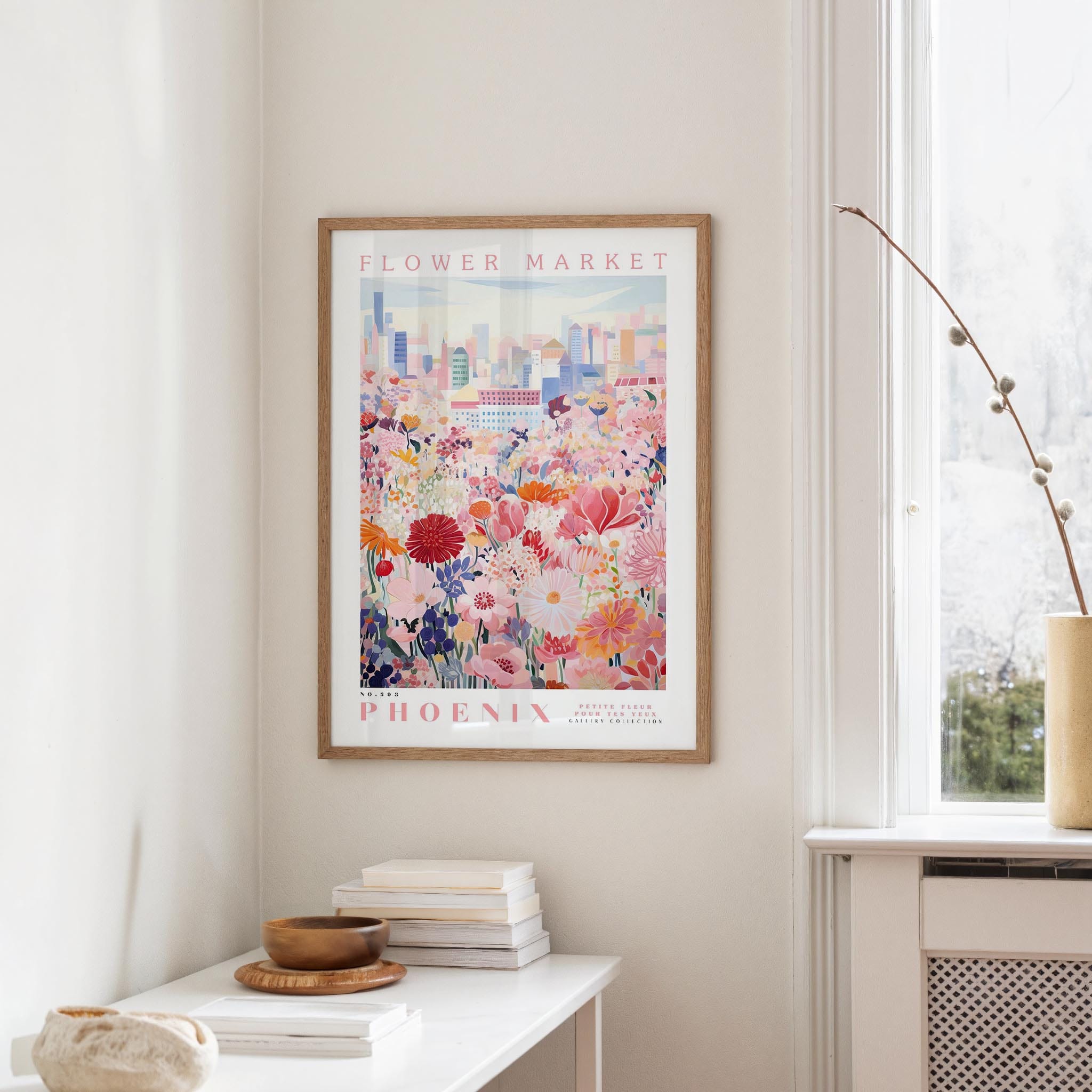 a framed poster of a colorful floral scene hanging on a wall, with a window and a small table with books and a vase in the foreground.