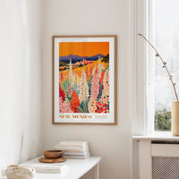 a framed poster of a colorful field of flowers hanging on a wall, with a white table and a vase of flowers in the foreground.