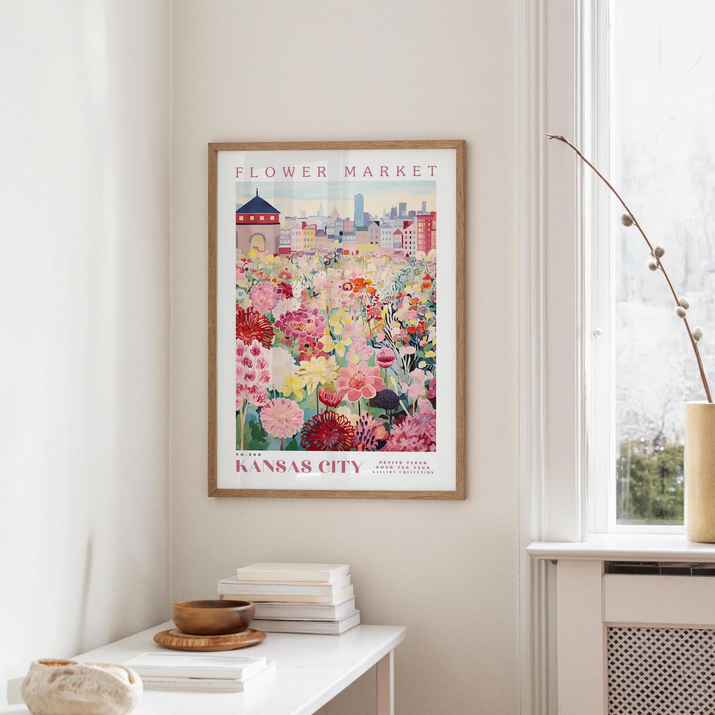 a framed poster of a colorful floral scene hanging on a wall, with a window and a small table with books and a vase in the foreground.