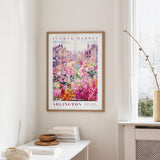 a framed poster of a colorful floral scene hanging on a wall, with a white table and a vase of flowers in the foreground.