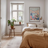a cozy bedroom with a large bed, a wooden nightstand, and a window with a plant on the sill. The room has a warm, inviting atmosphere with a neutral color palette and natural elements like plants and a rug.