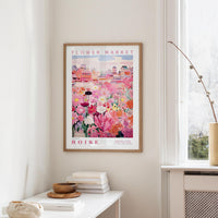 a framed poster of a colorful floral scene hanging on a wall, with a white table and a vase of flowers in the foreground.