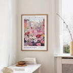 a framed poster of a colorful floral scene hanging on a wall, with a white table and a vase of flowers in the foreground.