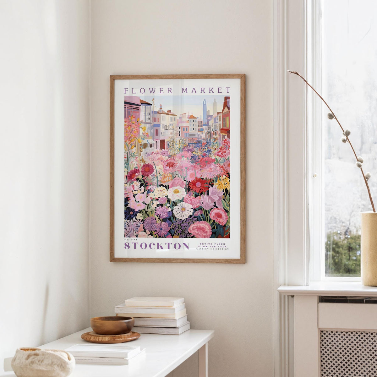 a framed poster of a colorful floral scene hanging on a wall, with a white table and a vase of flowers in the foreground.