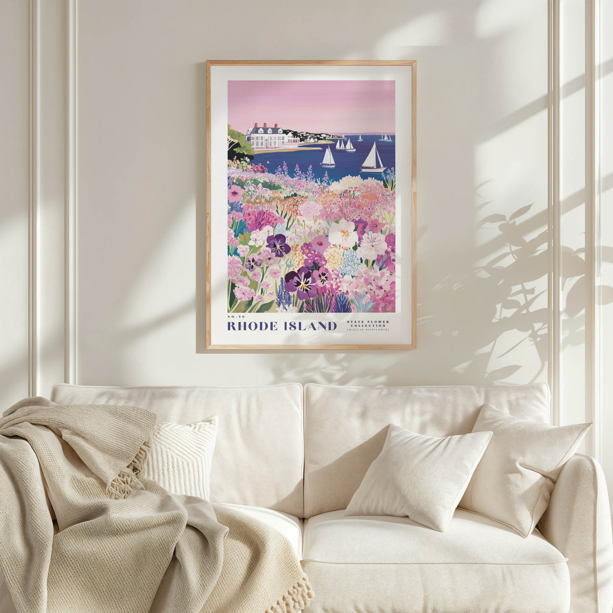a framed poster of a scenic view of Rhode Island, with a lush garden and sailboats in the background. The poster is hung on a wall above a white couch, creating a cozy and inviting atmosphere in the living room.