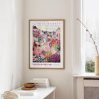 a framed poster of a colorful garden scene hanging on a wall, with a white table and a vase of flowers nearby.
