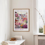 a framed poster of a colorful garden scene hanging on a wall, with a white table and a vase of flowers nearby.