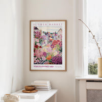 a framed poster of a colorful garden scene hanging on a wall, with a white table and a vase of flowers nearby.