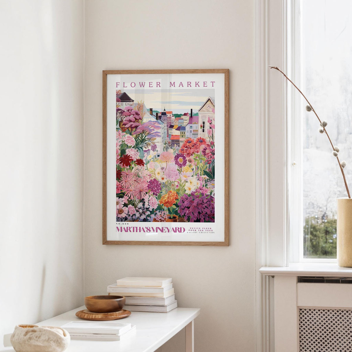 a framed poster of a colorful garden scene hanging on a wall, with a white table and a vase of flowers nearby.