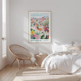 a bedroom with a bed, a wicker chair, and a framed poster of a flower market in Montauk.