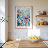 a kitchen scene with a framed poster of a colorful floral design hanging on the wall above a wooden countertop. On the countertop, there is a bowl of lemons and a plant. The kitchen also has shelves with various jars and containers, and a window letting in natural light.