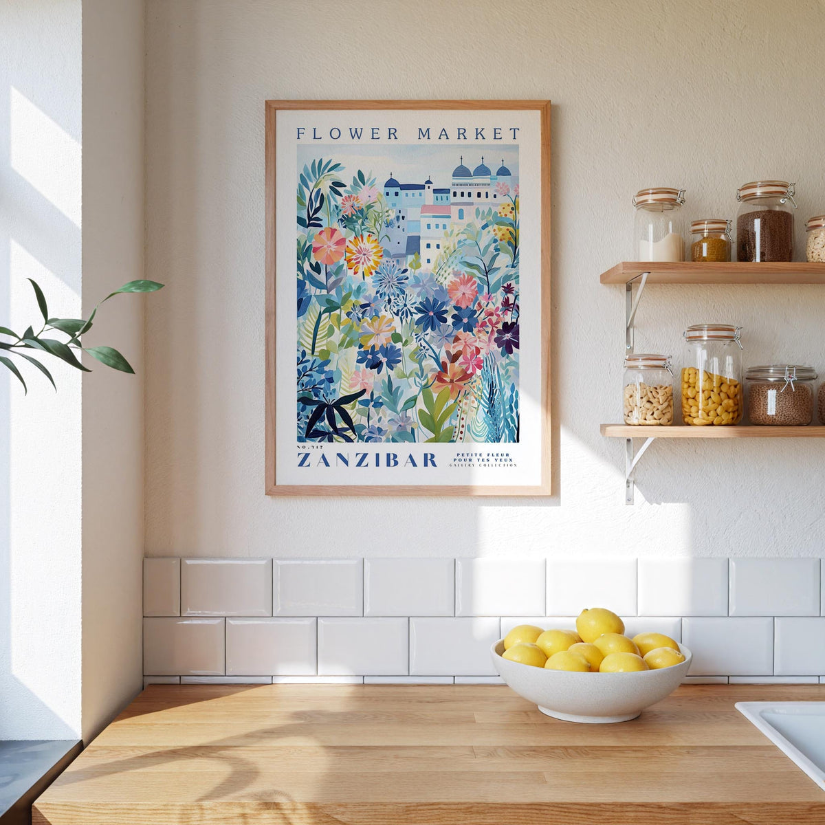 a kitchen scene with a framed poster of a colorful floral design hanging on the wall above a wooden countertop. On the countertop, there is a bowl of lemons and a plant. The kitchen also has shelves with various jars and containers, and a window letting in natural light.