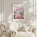 a framed poster of a colorful floral scene hanging on a wall above a white couch with pillows.
