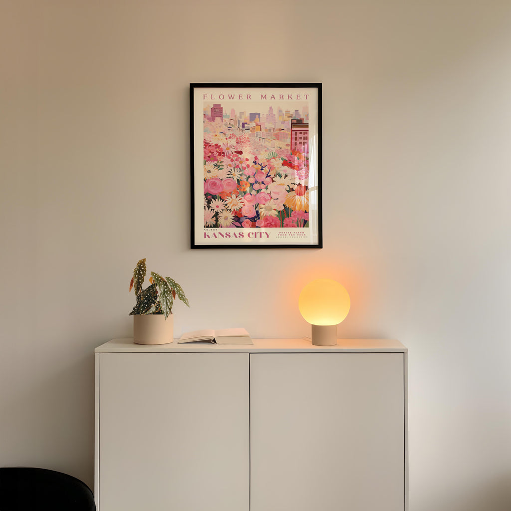a white cabinet with a potted plant and a lamp on top, set against a wall with a framed poster of a cityscape above it.