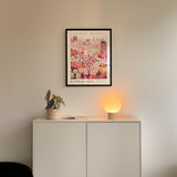 a white cabinet with a potted plant and a lamp on top, set against a wall with a framed poster of a cityscape above it.