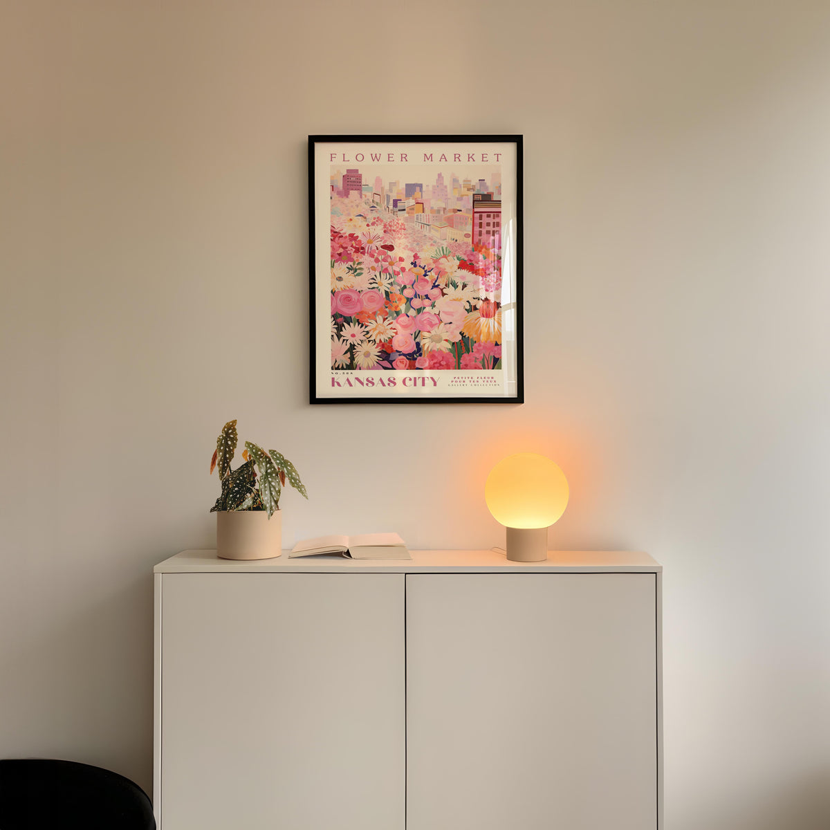 a white cabinet with a potted plant and a lamp on top, set against a wall with a framed poster of a cityscape above it.