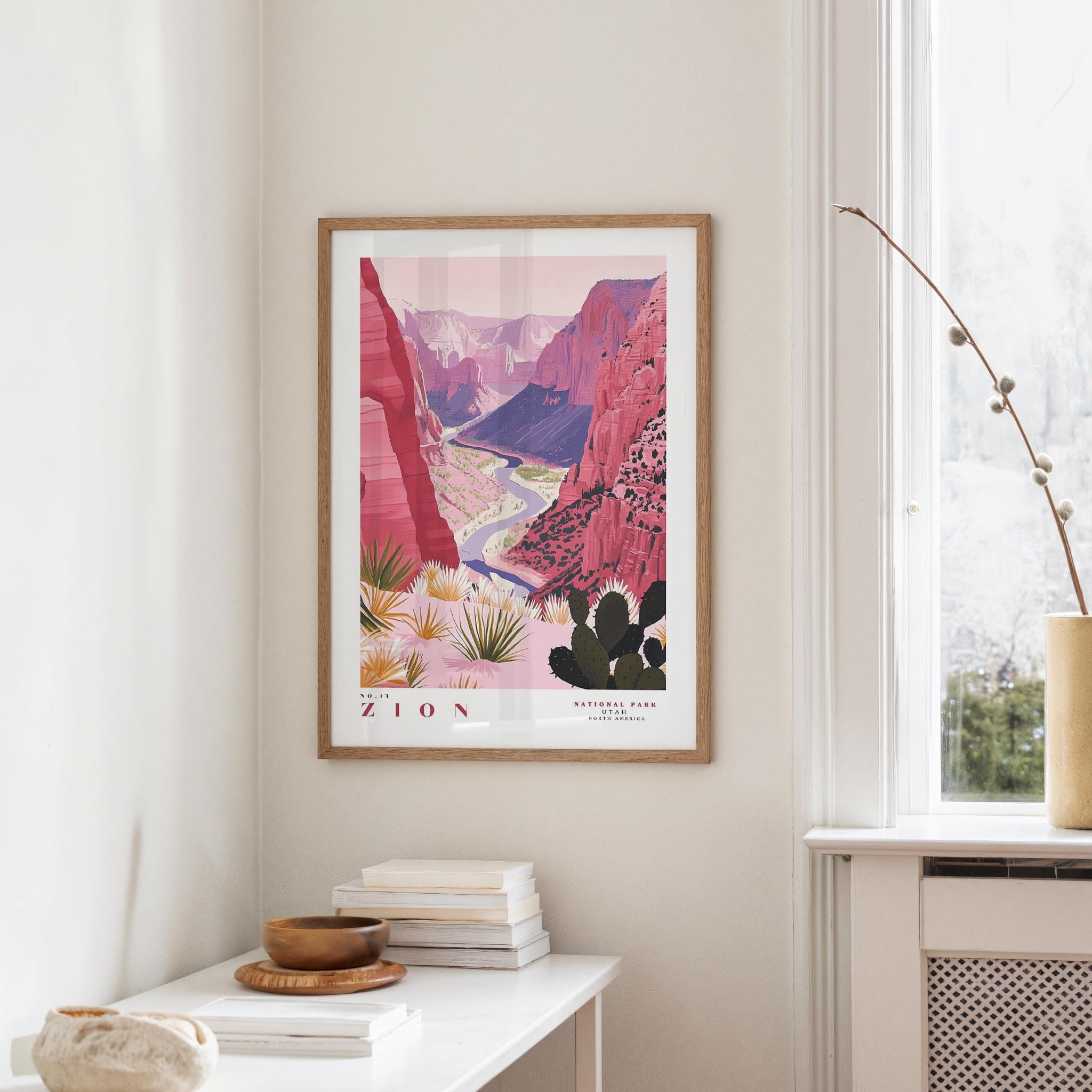 a framed poster of a mountainous landscape with a river, hanging on a wall in a room with a white table and a window.