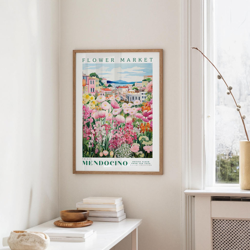 a framed poster of a colorful garden scene hanging on a wall, with a white table and a vase of flowers in the foreground.