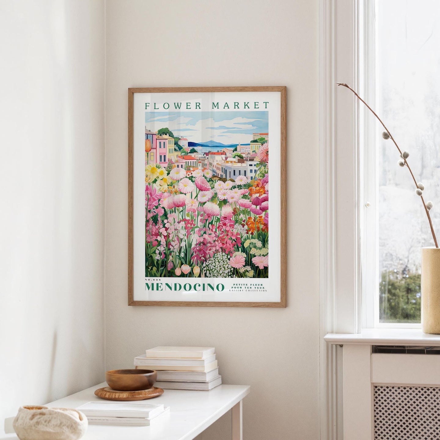 a framed poster of a colorful garden scene hanging on a wall, with a white table and a vase of flowers in the foreground.