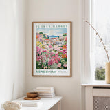 a framed poster of a colorful garden scene hanging on a wall, with a white table and a vase of flowers in the foreground.
