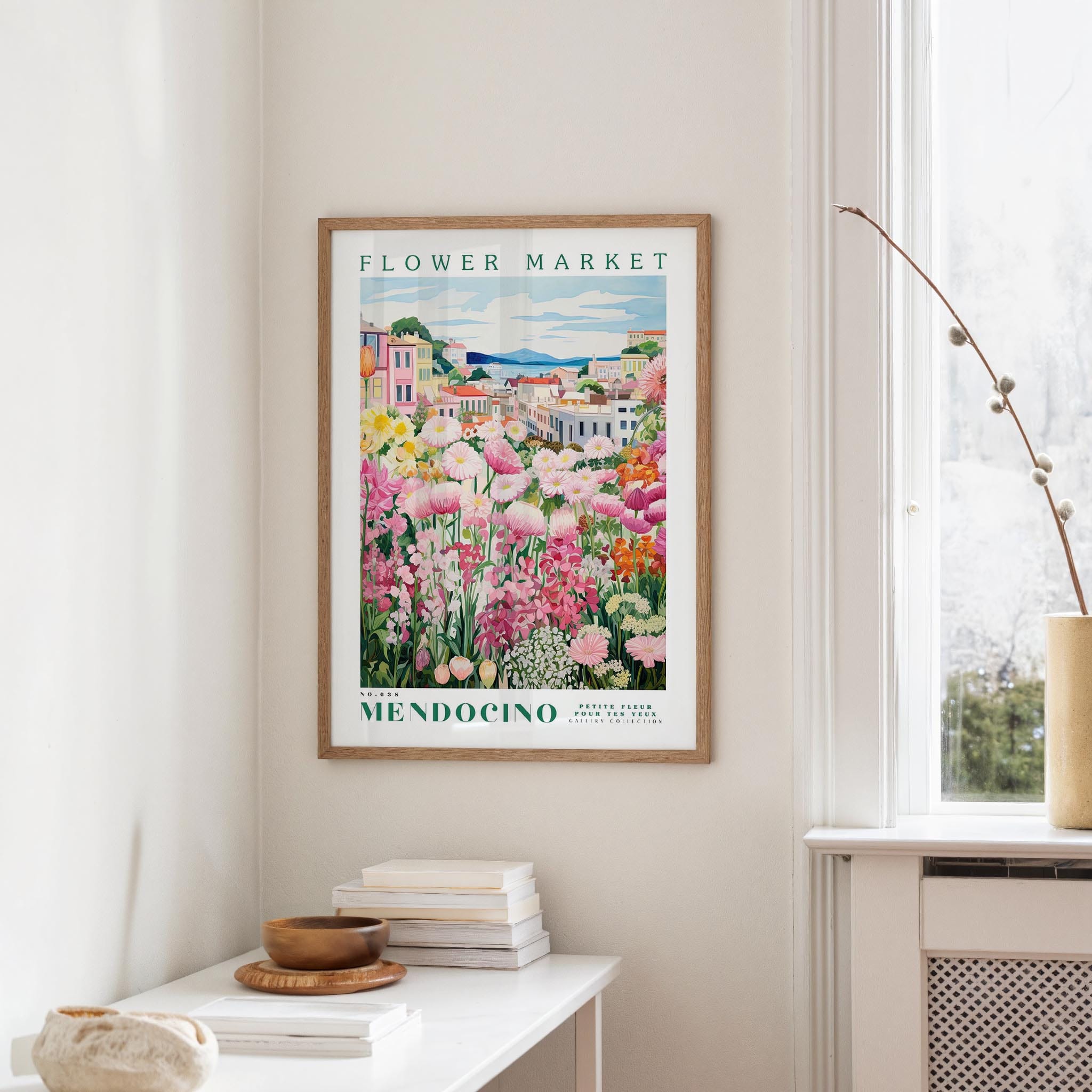 a framed poster of a colorful garden scene hanging on a wall, with a white table and a vase of flowers in the foreground.