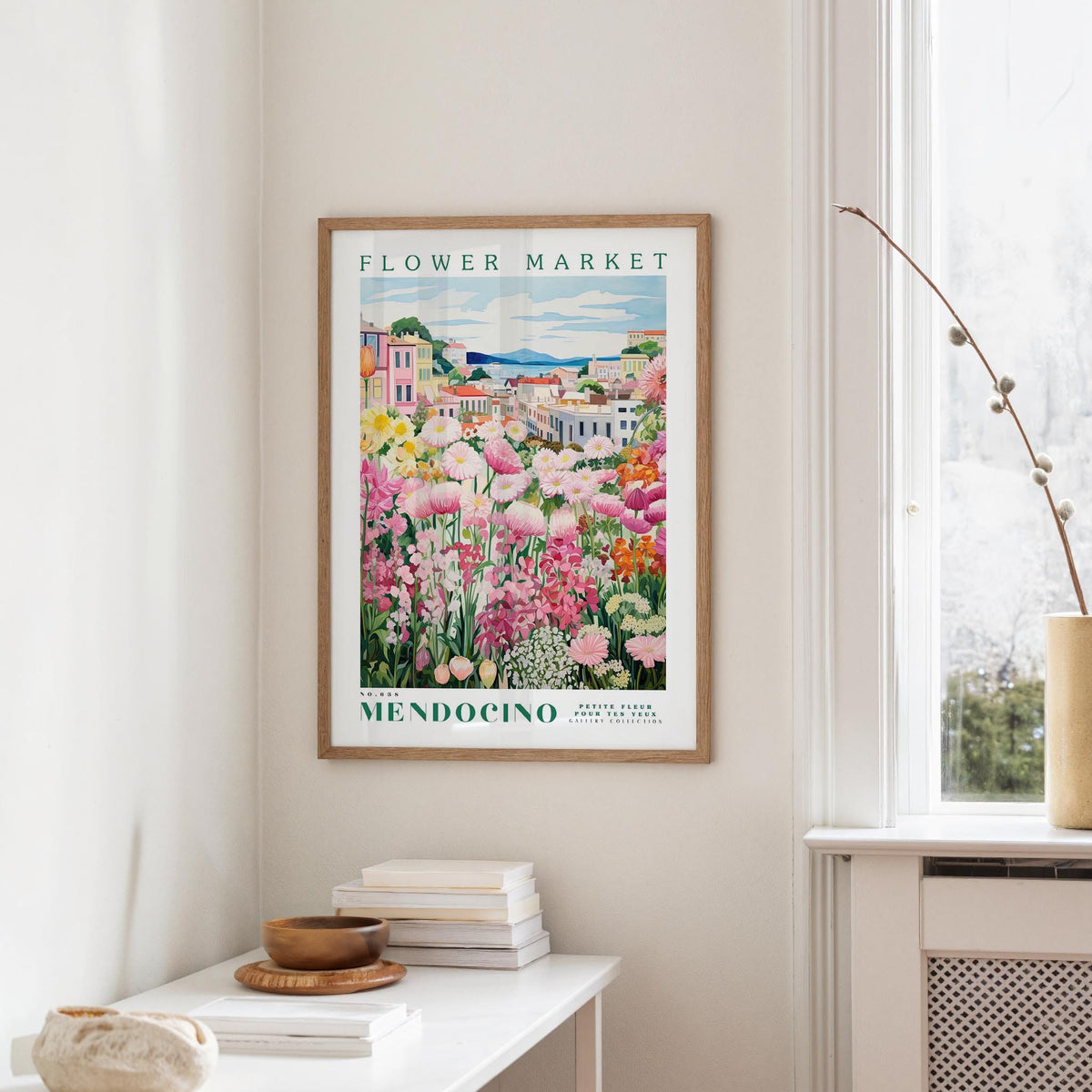 a framed poster of a colorful garden scene hanging on a wall, with a white table and a vase of flowers in the foreground.