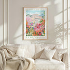 a framed poster of a colorful garden scene with flowers and buildings, hanging on a wall above a white couch.