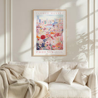 a framed poster of a cityscape with a field of flowers in the foreground, hanging on a wall above a white couch with a blanket draped over it.