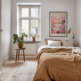 a cozy bedroom with a large bed, a wooden nightstand, and a window with a plant on the sill. The room has a neutral color scheme with a large poster of a flower market on the wall.