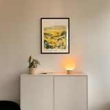 a white cabinet with a framed picture of a field hanging on the wall above it, and a small lamp on the cabinet.