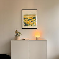 a white cabinet with a framed picture of a field hanging on the wall above it, and a small lamp on the cabinet.