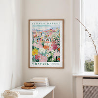 a framed poster of a colorful floral scene hanging on a wall, with a white table and a vase of flowers in the foreground.