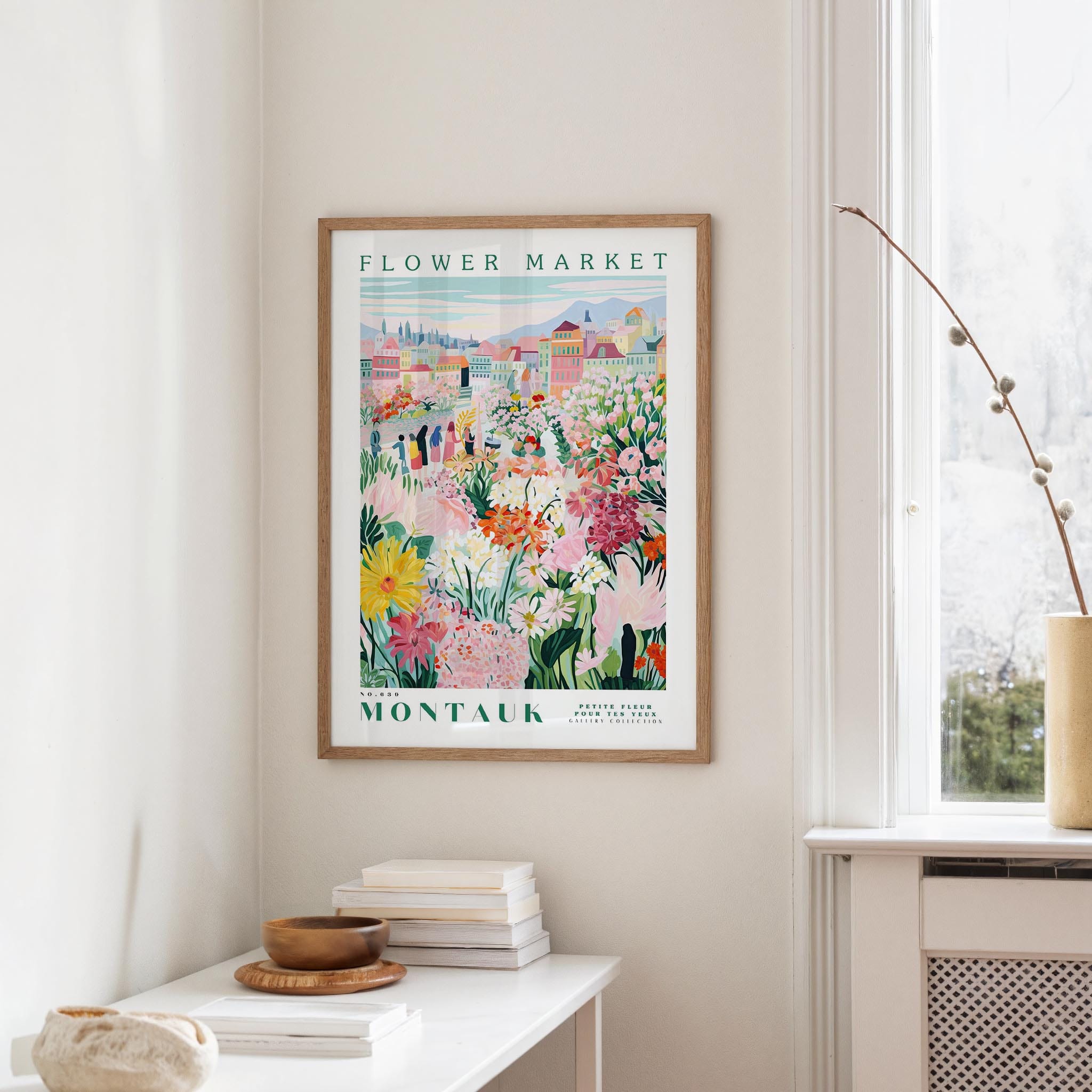 a framed poster of a colorful floral scene hanging on a wall, with a white table and a vase of flowers in the foreground.